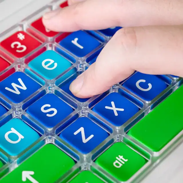 Close up image of a hand with a finger pressing a key on a colourful keyboard. The keyboard has a clear perspex keyguard separating the keys.