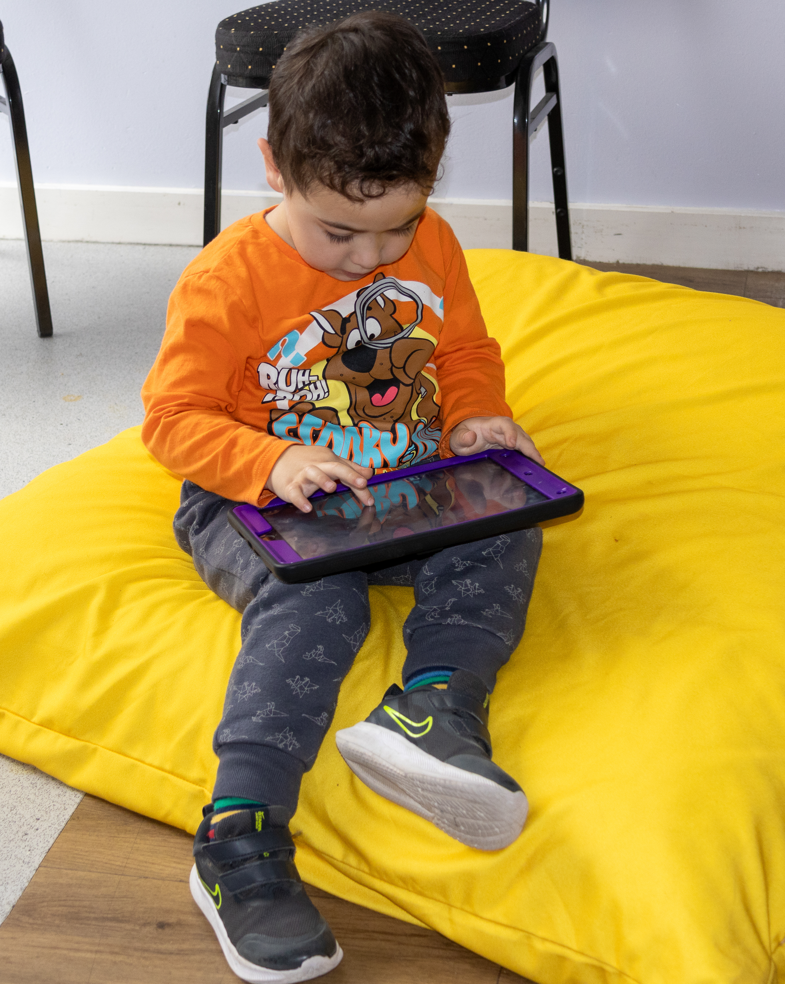 A young white boy with brown hair, wearing an orange top, sitting on a beanbag looking at an iPad and tapping the screen with his finger.