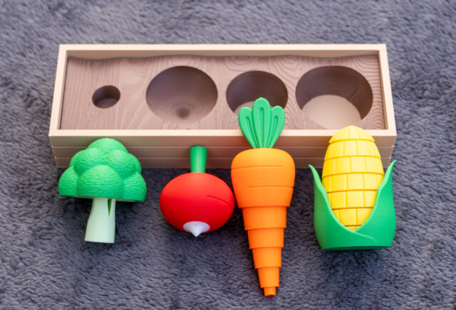 A small 3D printed raised garden bed with a broccoli, radish, carrot and corn sitting in front of the garden bed to display their function. The vegetables are fidget toys which provide an easy and accessible way to help self-regulate or concentrate by providing auditory, visual and/or tactile input.