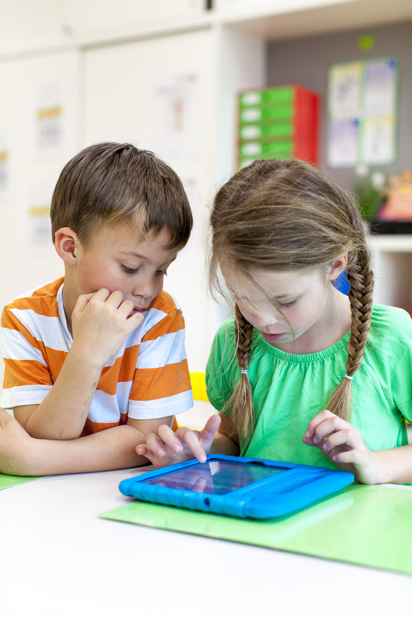 A boy in an orange and white striped t-shirt is sitting next to a girl with her hair in plaits, wearing a green t-shirt in a classroom. They are looking at an iPad on the table.