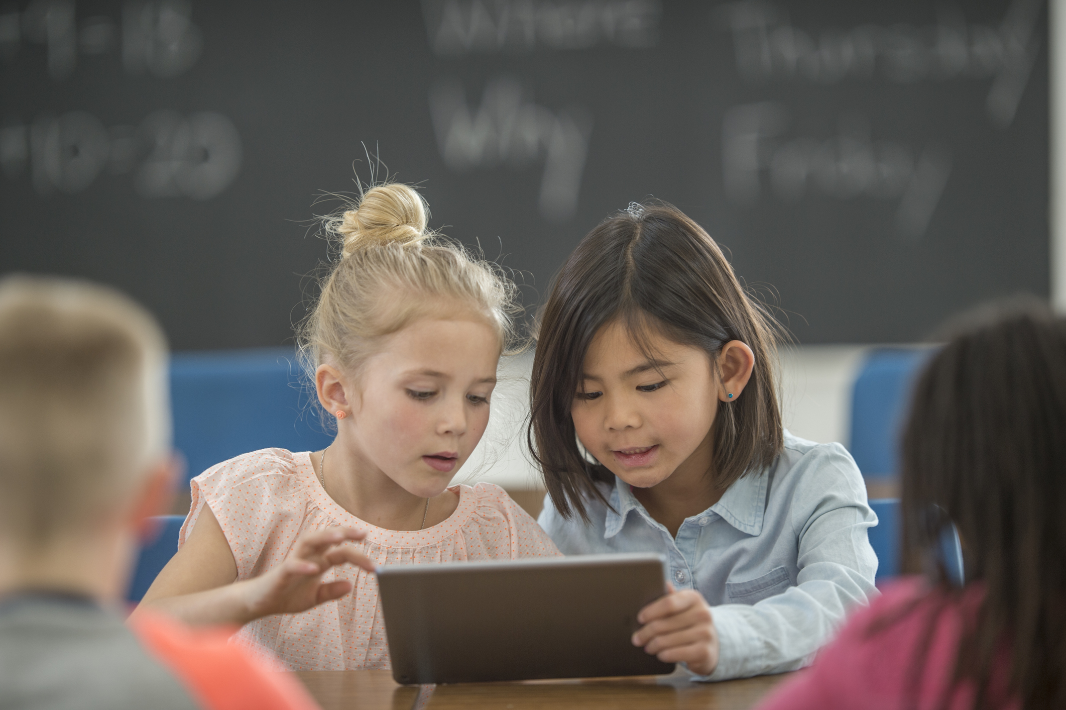 A young white girl and a young Asian girl looking at a tablet together. They are in a classroom.