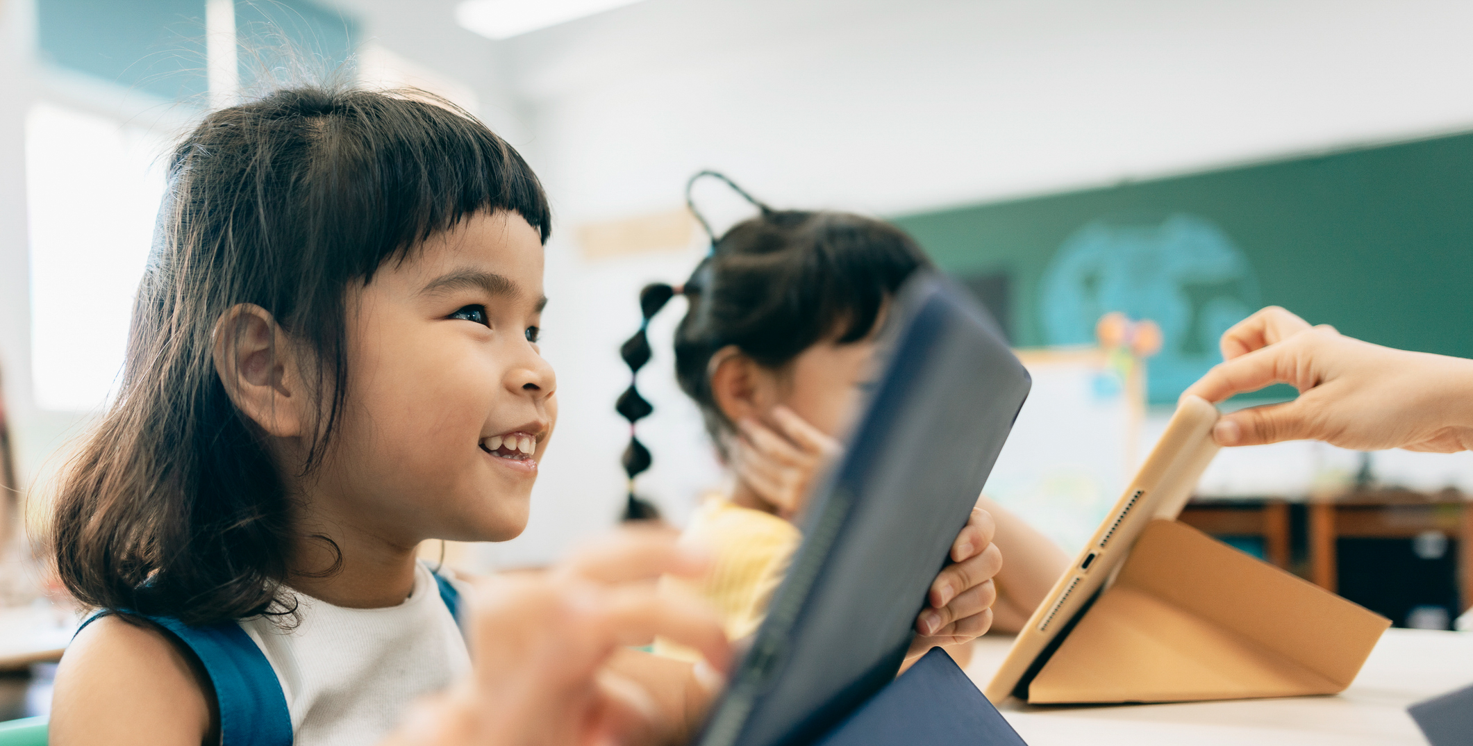 A young asian girl in a classroom, looking at a tablet and smiling. She is tapping the iPad with her finger.