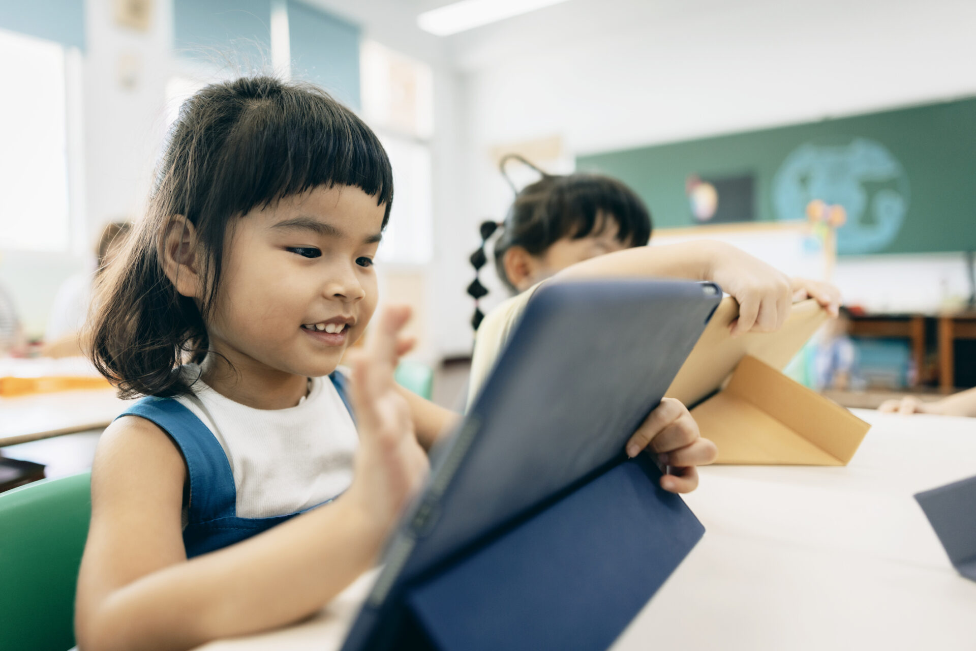 A young asian girl in a classroom, looking at a tablet and smiling. She is tapping the iPad with her finger.