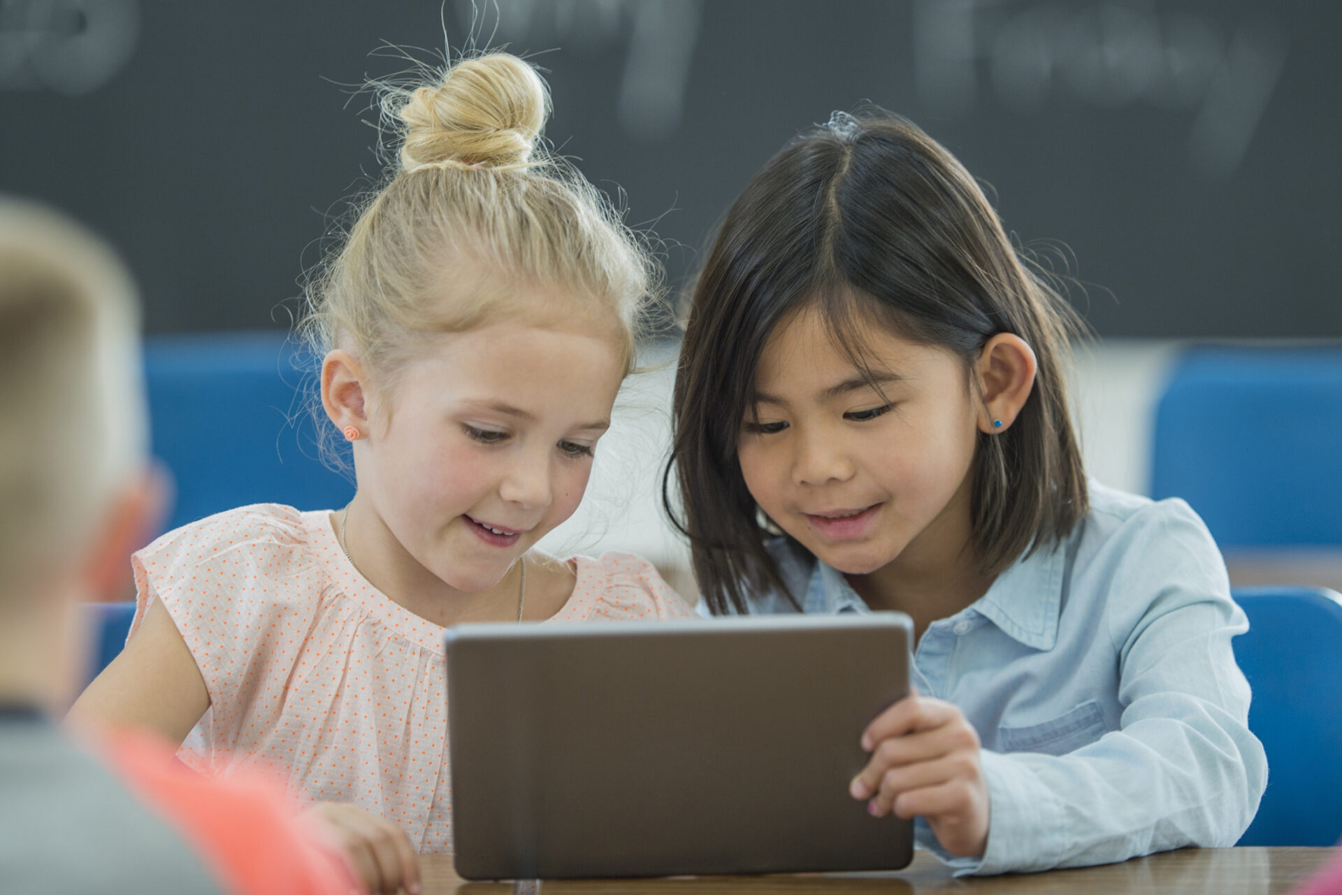 Two school aged children look at a digital tablet together. A young blonde white girl is wearing a pink top and a young Asian dark haired girl is wearing a blue button-up blouse.