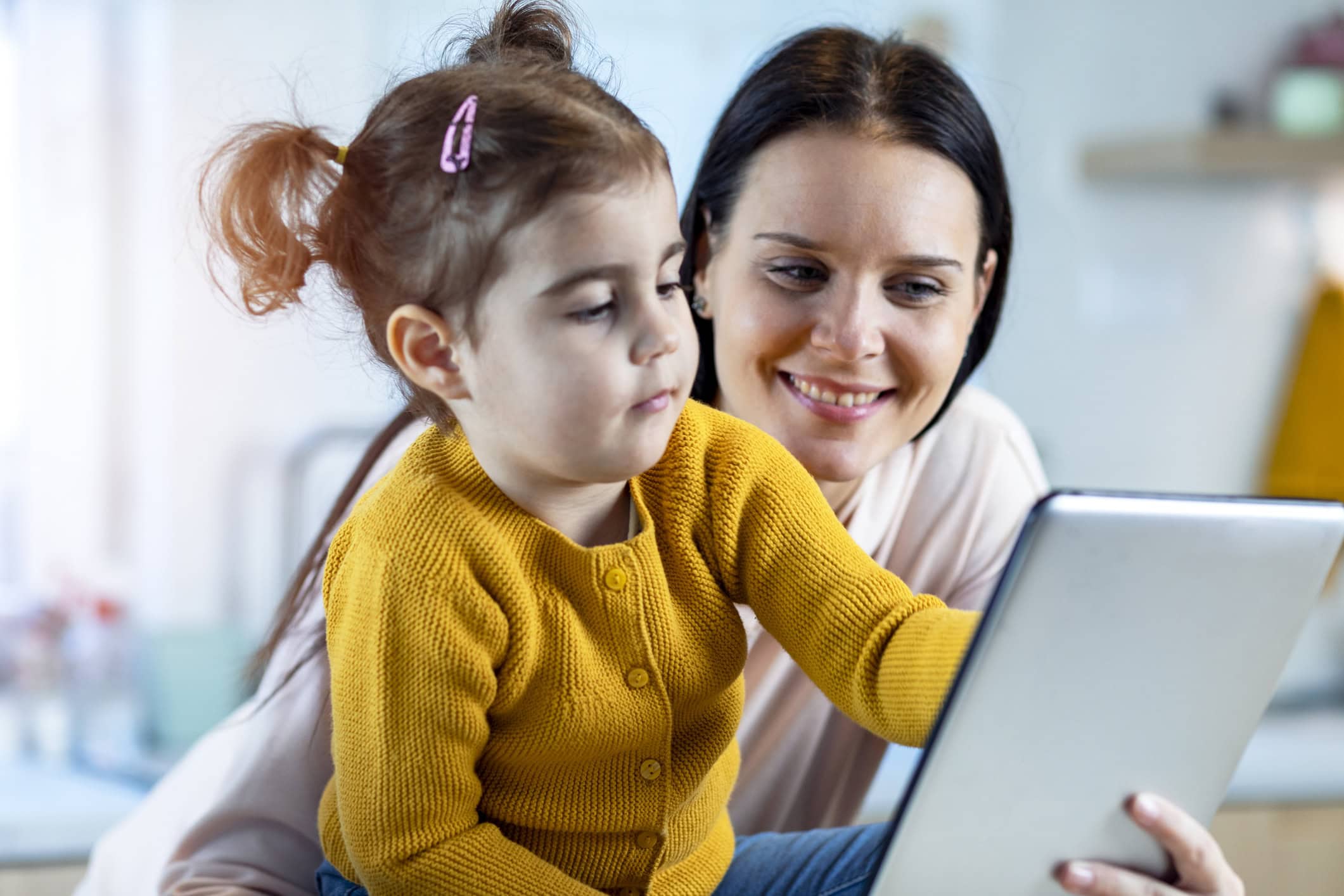 A young child with pigtails wearing a yellow cardigan, leaning forward to touch an iPad. She is sitting with a white dark haired female adult.