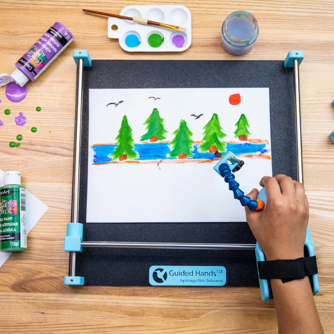 A top view of Guided Hands assistive technology device with a piece of paper fixed to the bed which has drawings of trees. A white person's hand is placed in the handpiece which has a marker in the handpiece.