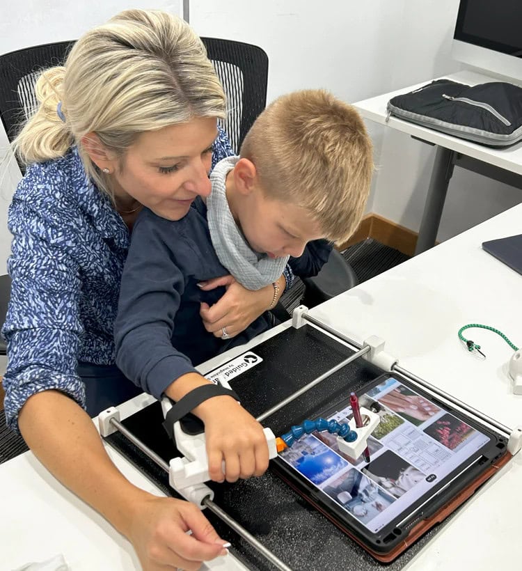 Young white blond haired boy sitting on a white adult female's lap at a table, using Guided Hands assistive technology device.