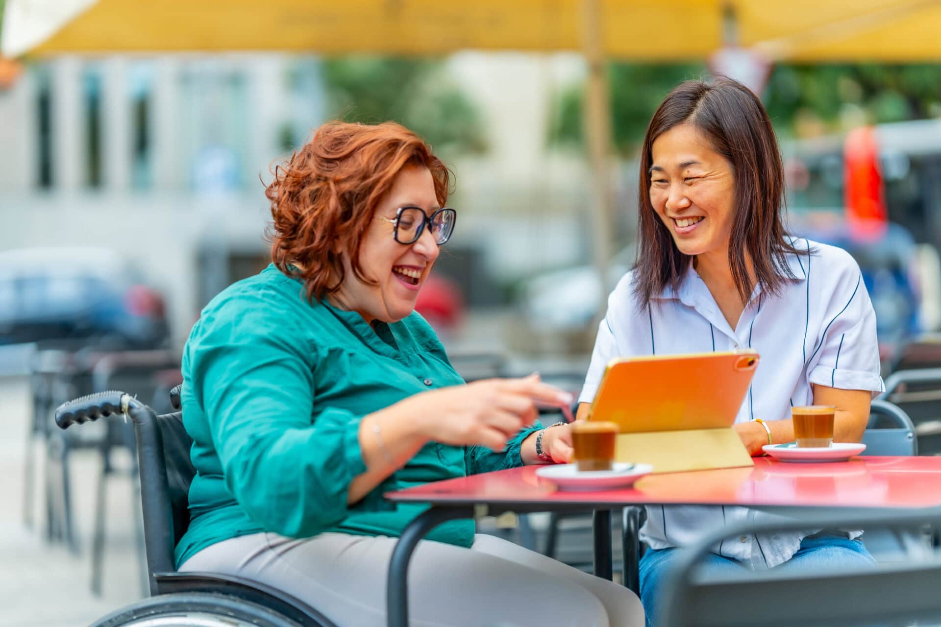 A white woman and an Asian woman at a coffee shop. They are sitting at a table chatting with an iPad in front of them.
