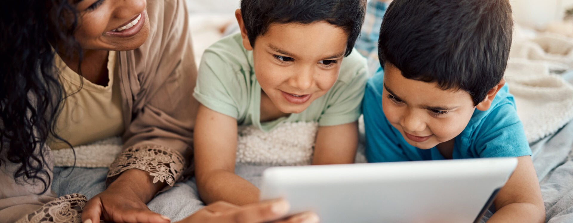 Two young boys looking at an iPad together with their mum. They are sitting on the floor together lying on their stomachs.