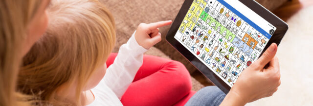 Child using an iPad with an AAC communication app (symbol-based grid) while interacting with an adult, supporting speech and communication development.
