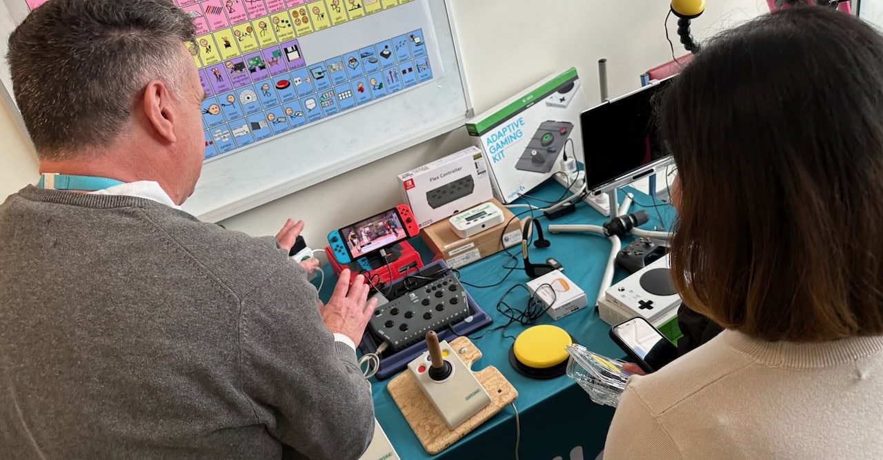 Two people sit at a table exploring a range of assistive technology and adaptive gaming equipment. Visible on the table are a Nintendo Switch, adaptive gaming controller, joystick, large yellow switch, switch interfaces, and other access devices. In the background, a communication display with AAC symbols is mounted on a wall, highlighting a consultation or demonstration focused on accessible gaming and communication technology.