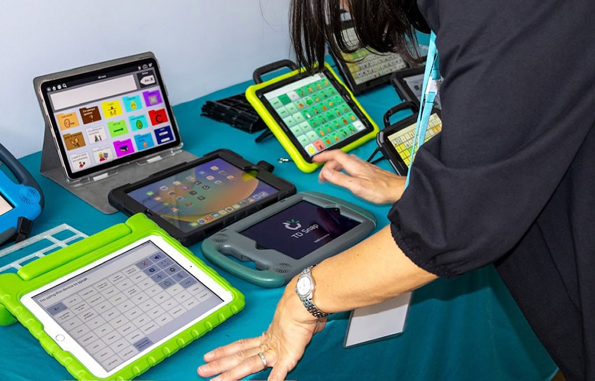 A person explores a range of AAC and communication devices displayed on a table, including iPads with communication apps, dedicated speech-generating devices, and tablet-based AAC systems used for assistive communication assessment and trials.