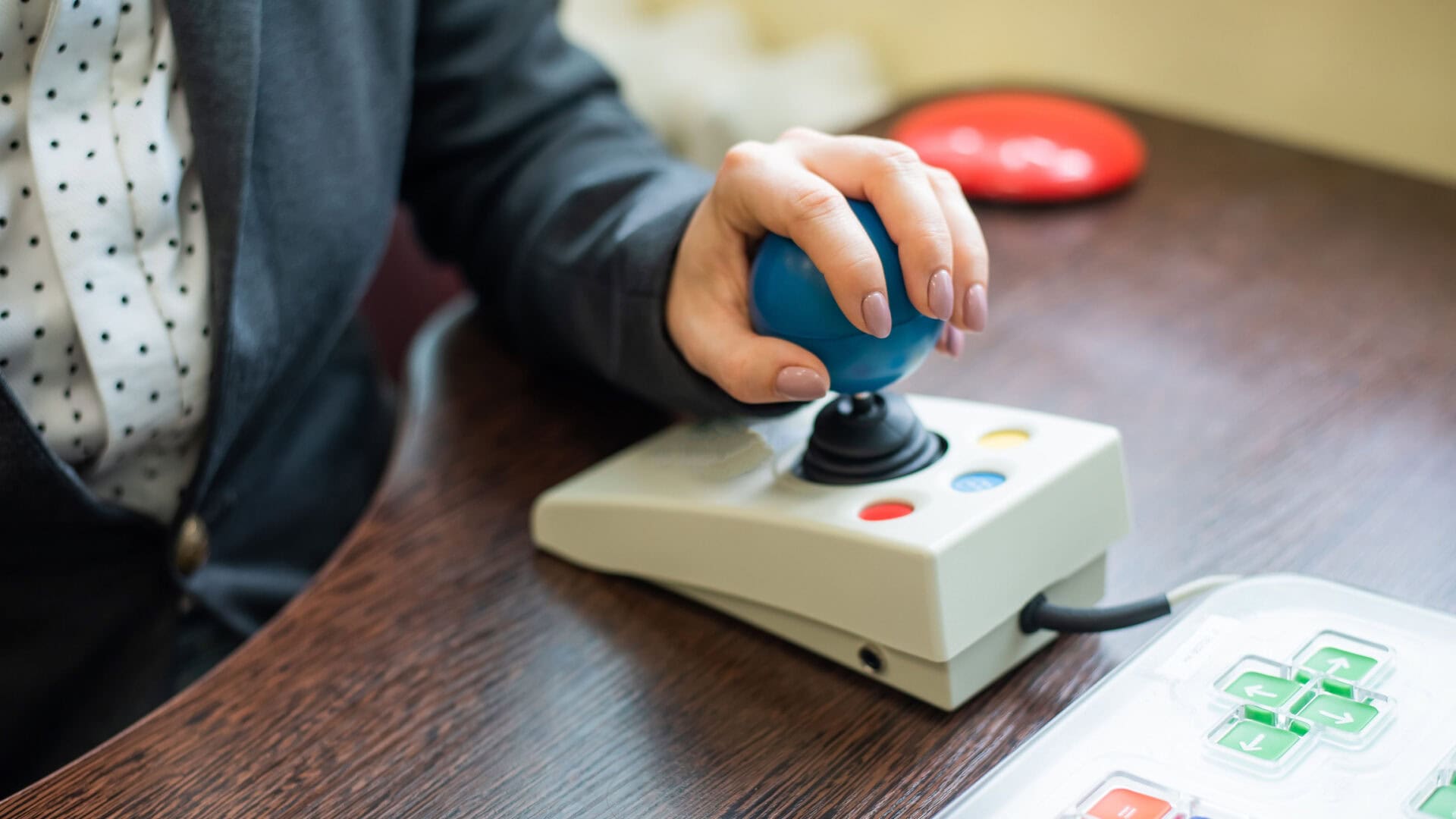 Close-up of a person using a blue joystick-style assistive technology controller mounted on a tabletop device. The person's hand rests on the joystick while a control panel with directional arrow buttons is visible nearby, demonstrating accessible computer or wheelchair control technology.