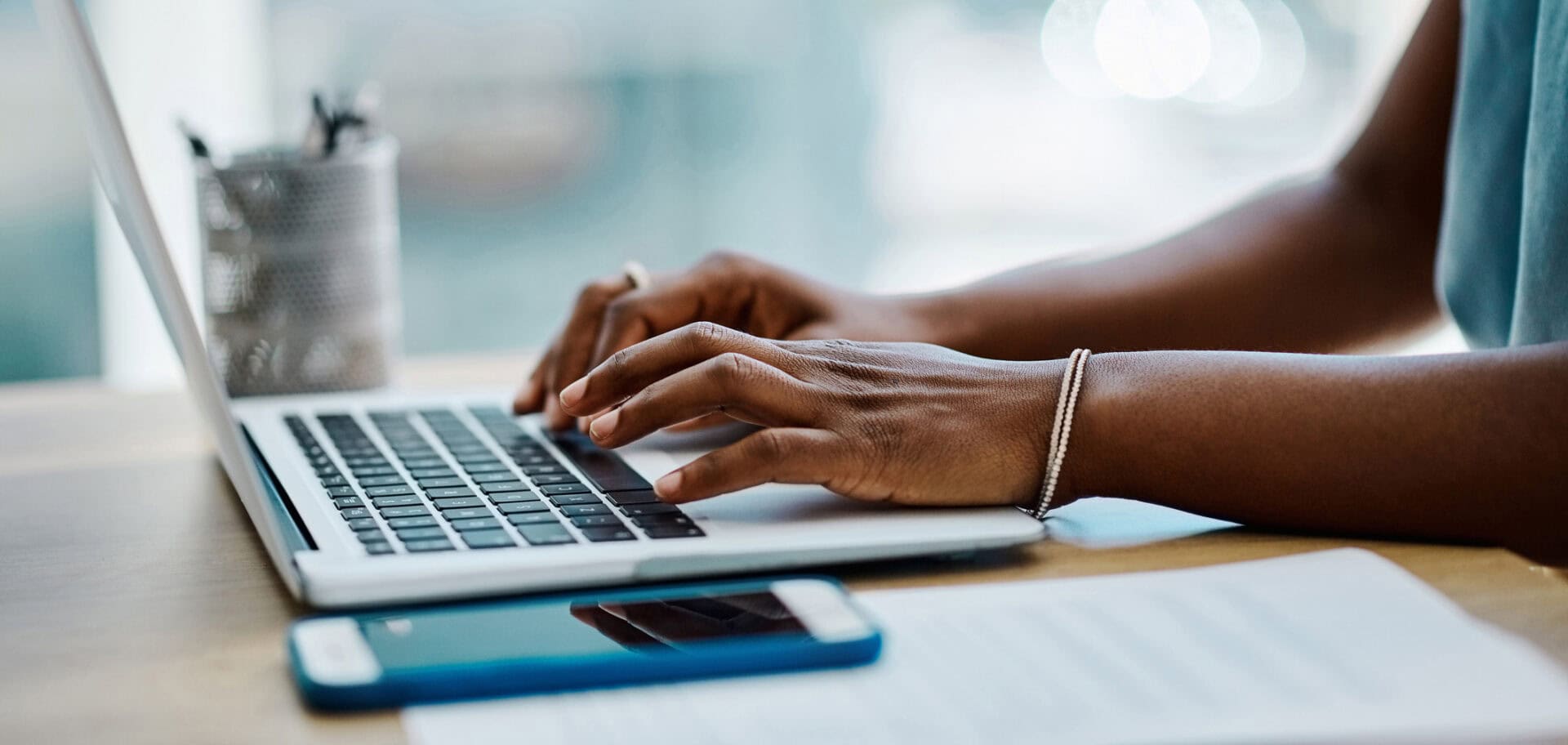 Close-up of a person typing on a laptop at a desk. A smartphone lies in the foreground beside a notebook, while a pen holder sits in the background. The image has a bright, modern workspace feel with a softly blurred background.