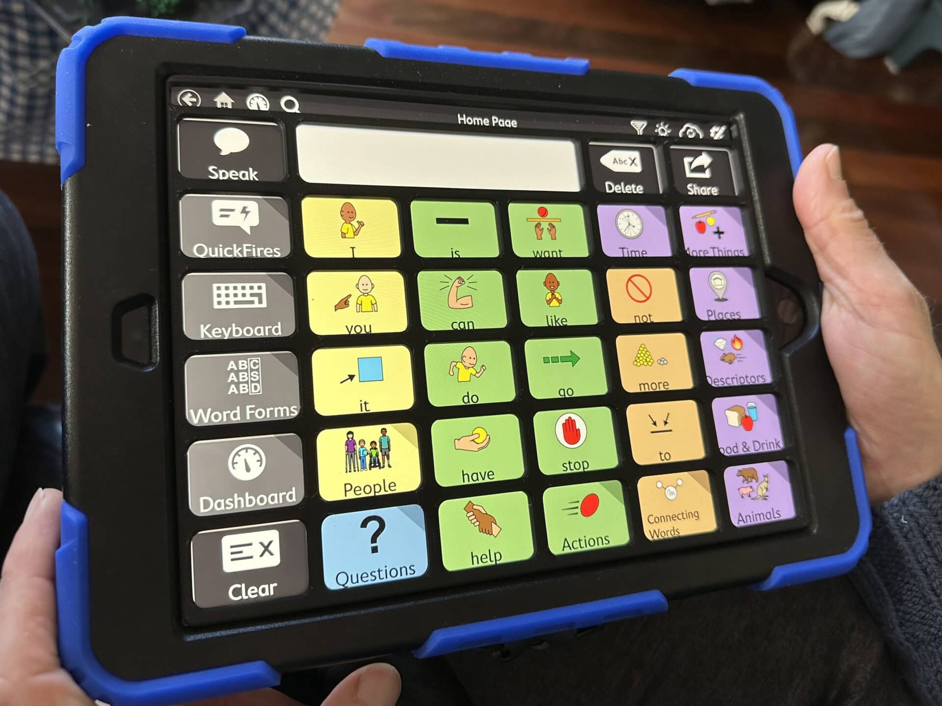 An iPad in a MacGuy Sumo case displaying the AAC app TD Snap with a Motor Plan 30 page set. There is a person's hands holding the iPad and there is a black keyguard fitted to the iPad screen.